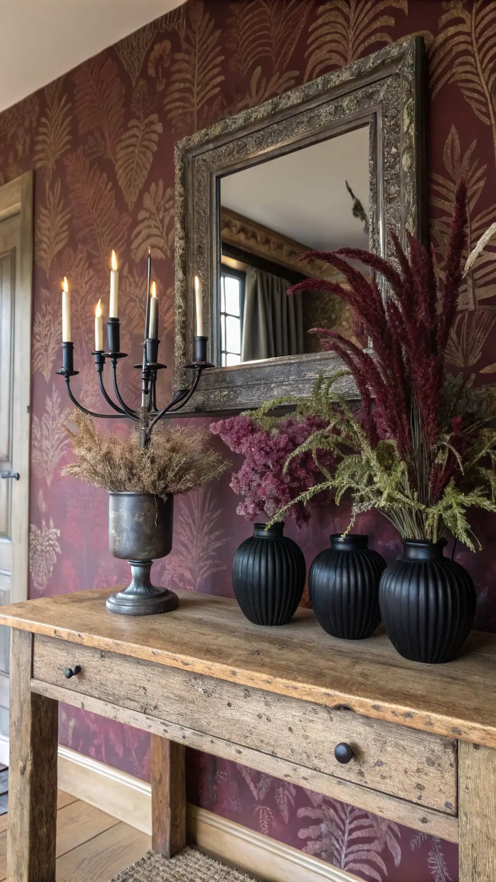 Antique oak console table set against burgundy Morris & Co wallpaper, adorned with dried purple amaranthus and ferns in matte black vases, reflected in a tarnished silver mirror illuminated by candlelight.